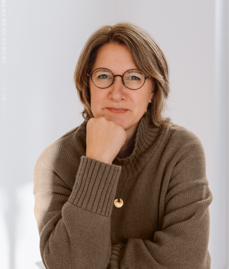 Nicole sitz an einem Tisch, ihr Kopf aufgestützt auf ihrer Hand und lächelt in die Kamera
